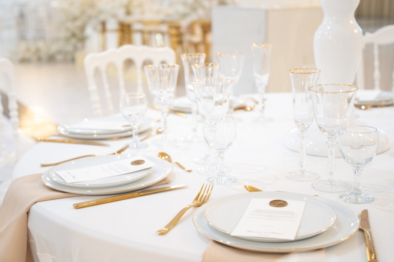 Table élégamment dressée avec nappes blanches, couverts dorés, verres à pied et menus posés sur les assiettes.