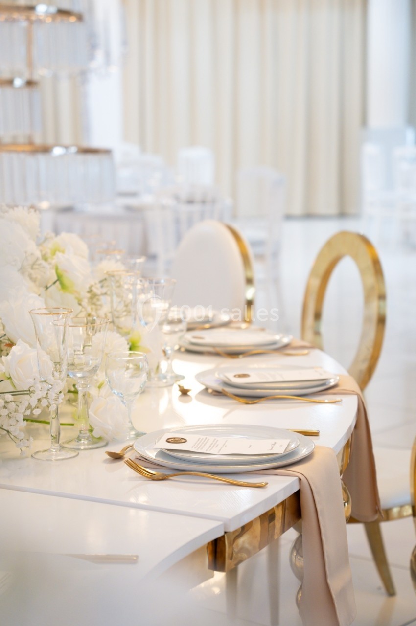 Table élégamment dressée avec vaisselle blanche, couverts dorés et décoration florale blanche dans une salle lumineuse.