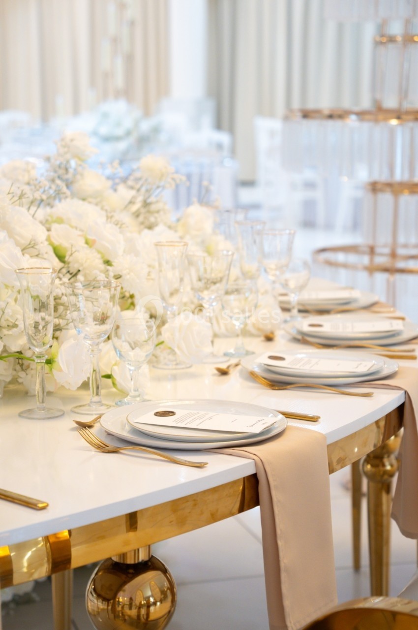 Table élégamment dressée avec vaisselle blanche, couverts dorés et décoration florale blanche dans une salle lumineuse.