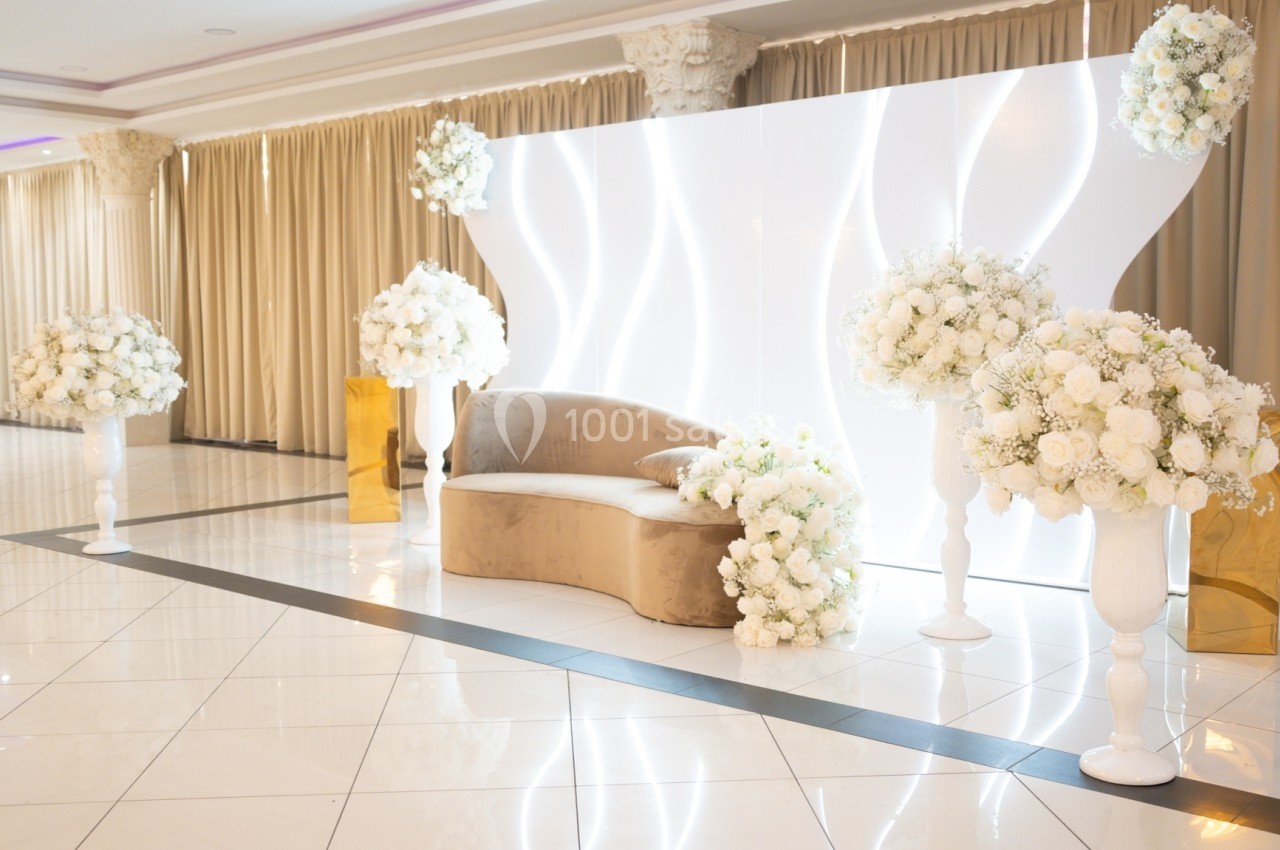 Canapé beige entouré de bouquets de fleurs blanches, devant un mur lumineux ondulé, dans une salle élégante.