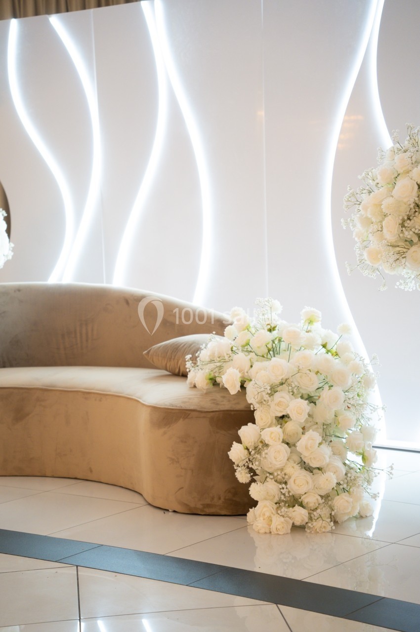 Canapé beige avec coussin, décoré de roses blanches, devant un mur lumineux aux motifs ondulés.