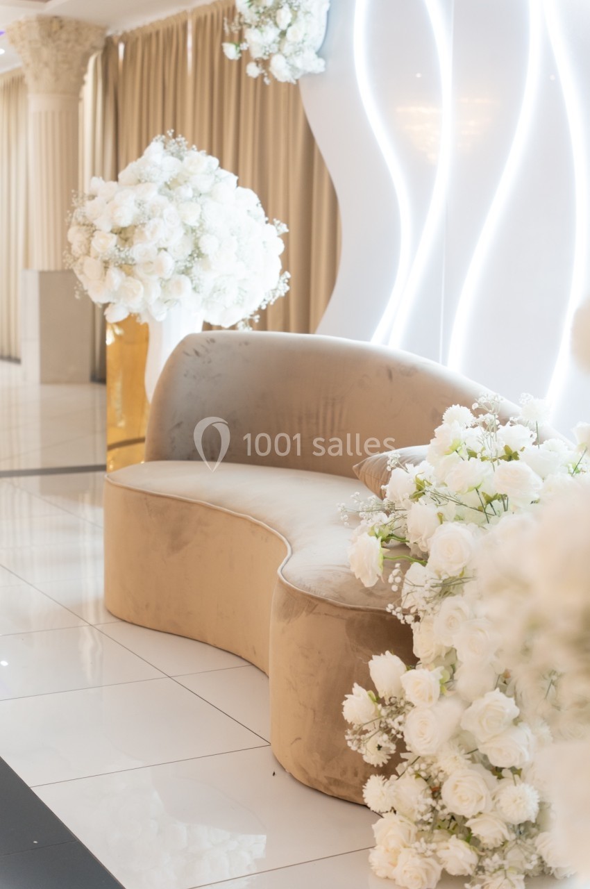 Canapé beige entouré de bouquets de fleurs blanches, dans un espace lumineux avec rideaux et décorations modernes.