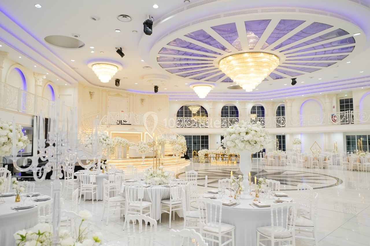 Salle de réception élégante avec tables décorées de fleurs blanches, lustre central imposant et éclairage lumineux.