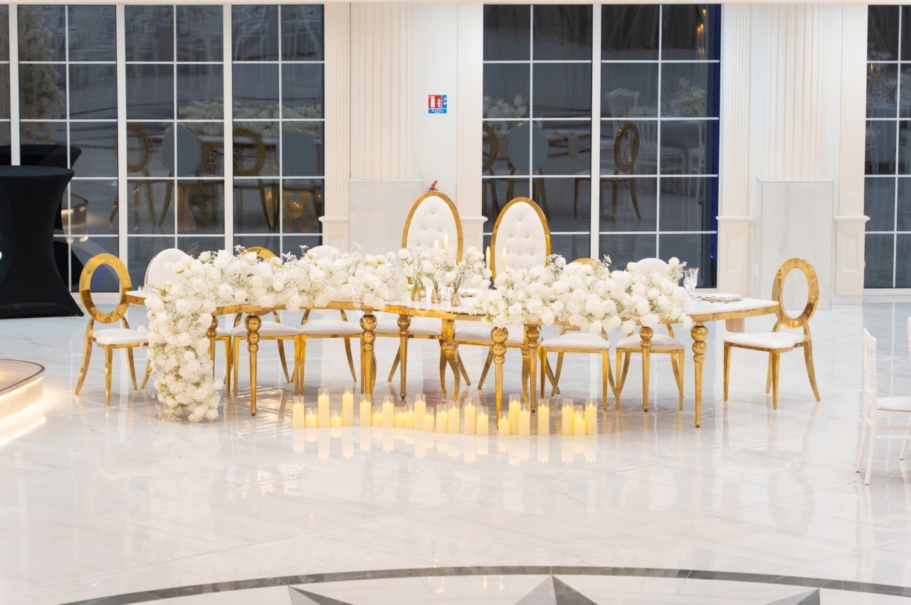 Table décorée avec des fleurs blanches, chaises dorées et bougies au sol dans une salle lumineuse avec grandes fenêtres.