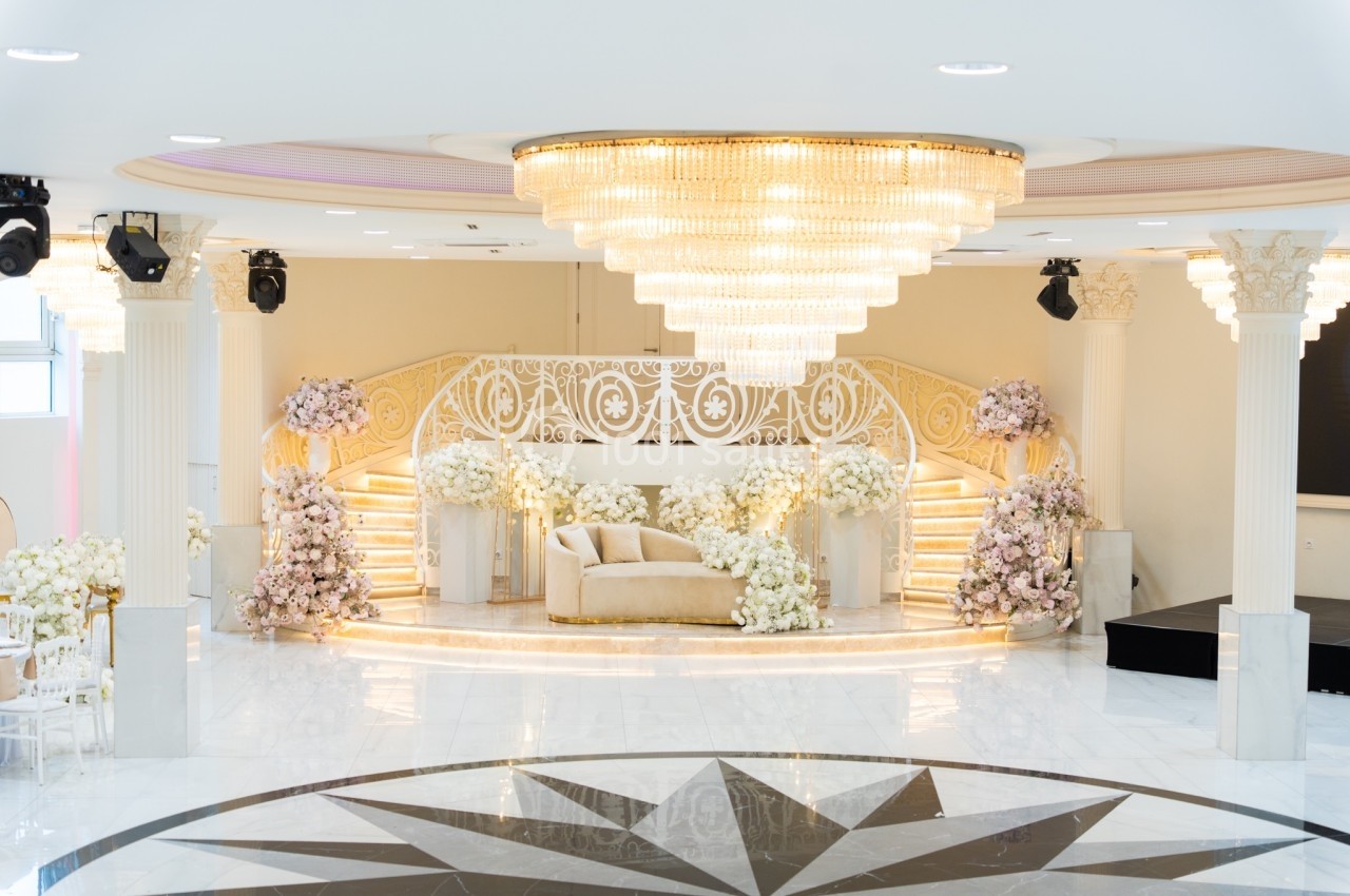 Salle de réception élégante avec escalier orné, canapé beige et décor floral abondant sous un grand lustre.