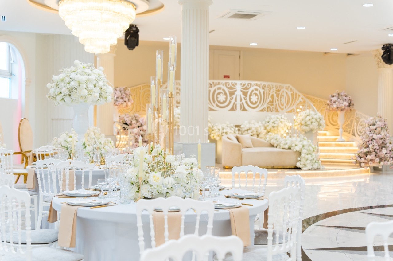 Salle de réception élégante avec tables décorées de fleurs blanches et escalier orné de motifs raffinés.