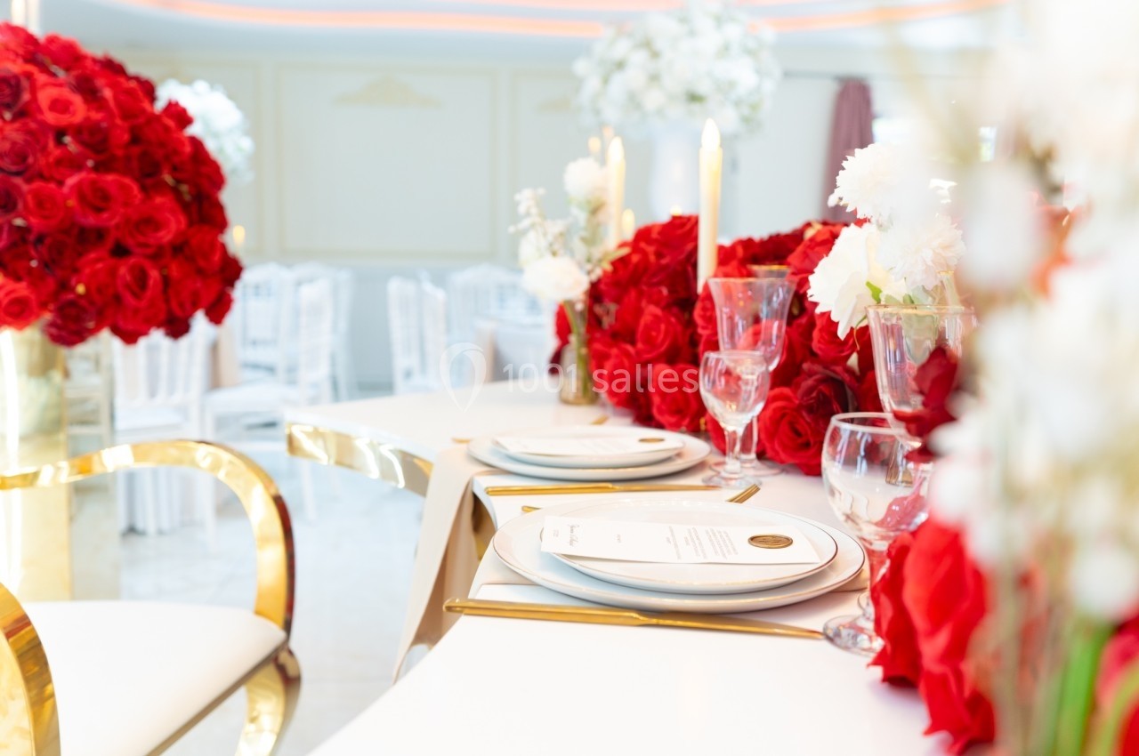 Table élégamment dressée avec des assiettes, des verres et des bouquets de roses rouges dans une salle lumineuse.