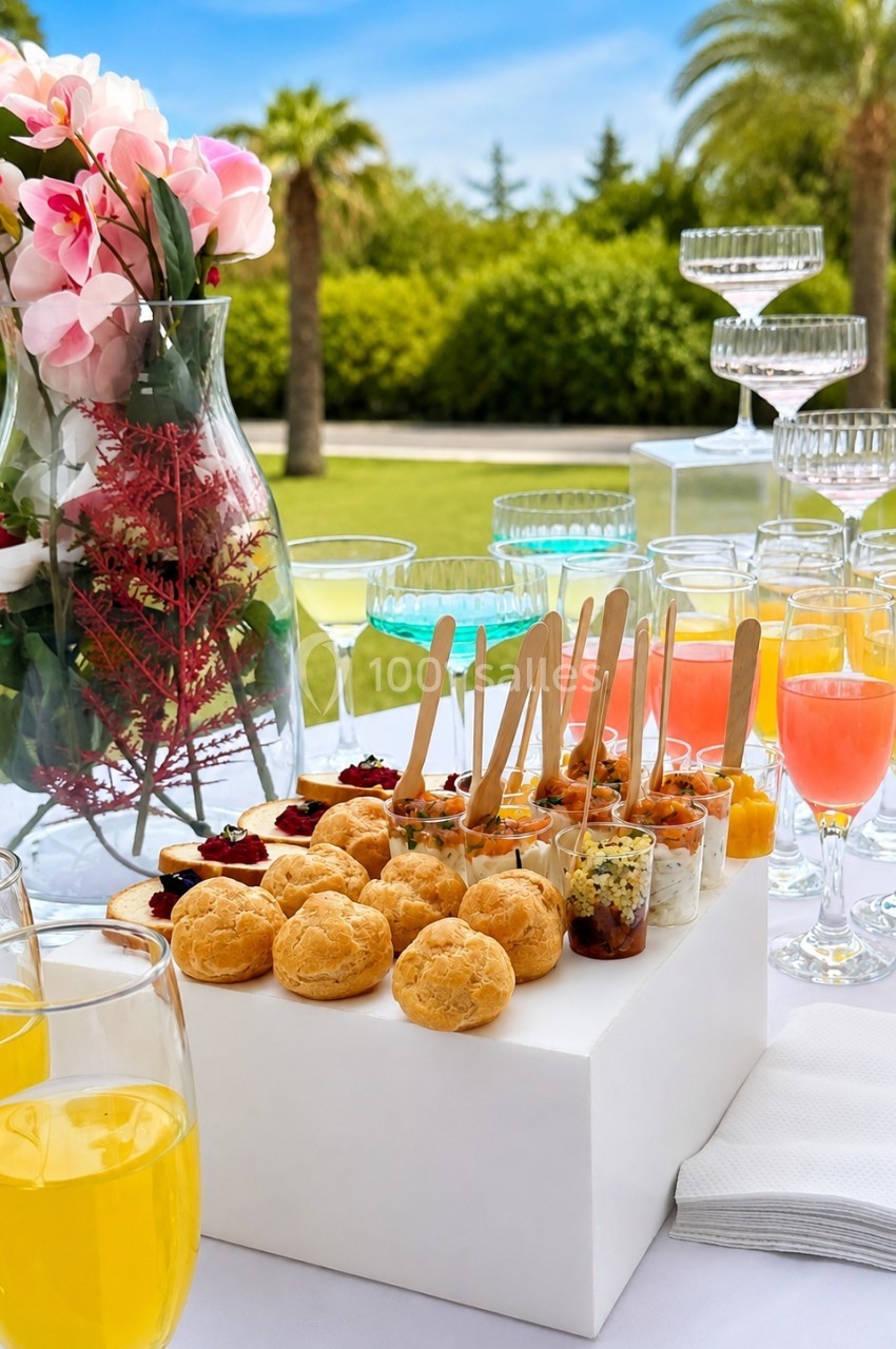 Buffet en plein air avec verrines, choux salés, cocktails colorés et vase de fleurs, sur fond de jardin verdoyant.