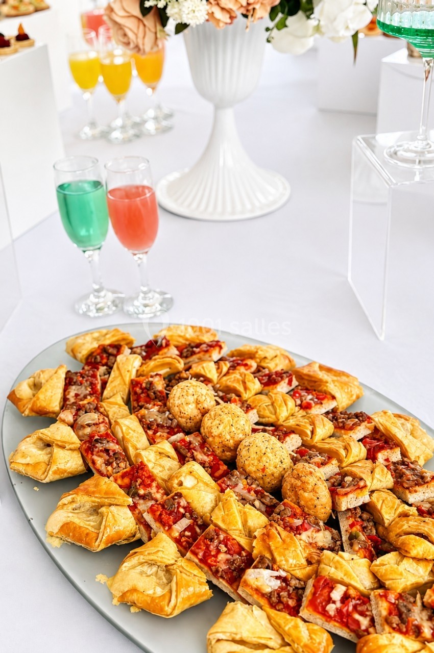 Plateau de pâtisseries salées variées sur une table blanche, avec des verres de cocktails colorés en arrière-plan.