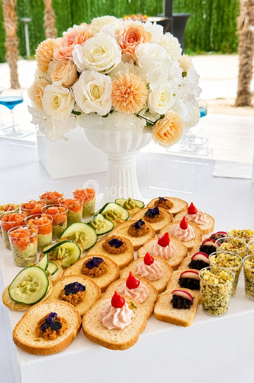 Plateau de canapés variés avec verrines, toasts garnis et bouquet de fleurs en arrière-plan.