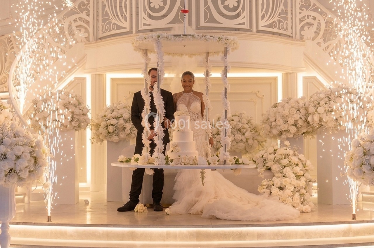 Un couple en tenue de mariage se tient devant un gâteau à plusieurs étages dans une salle décorée de fleurs blanches.
