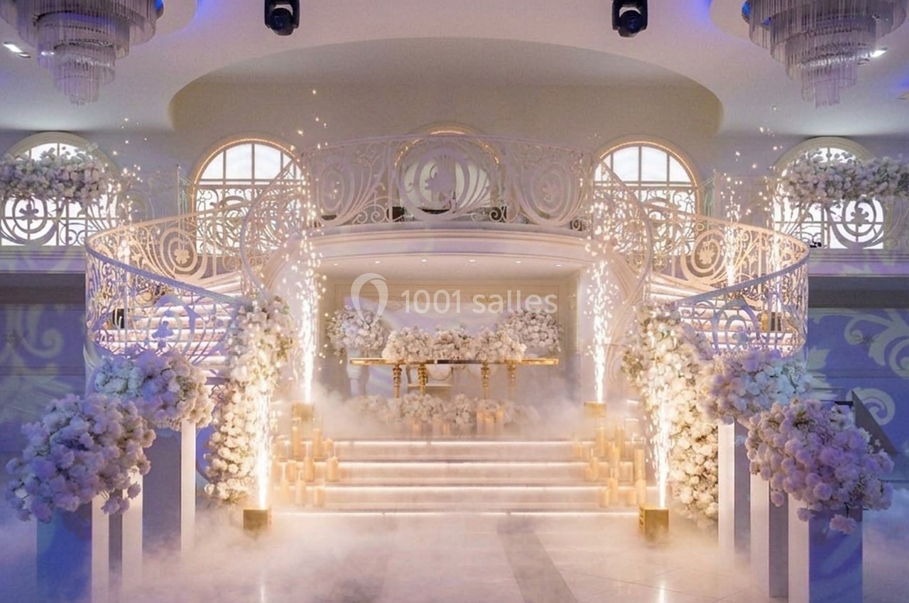 Salle de réception décorée pour un mariage avec des fleurs blanches, des escaliers ornés et des effets lumineux.