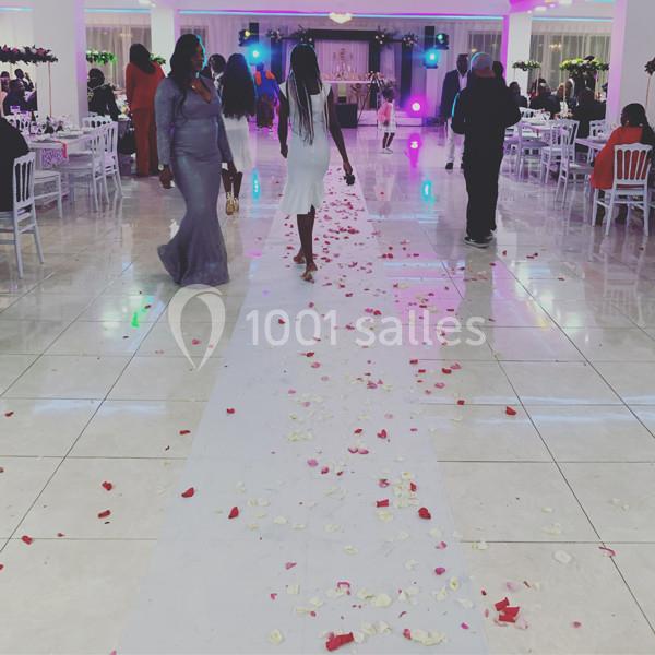 Allée recouverte de pétales de fleurs dans une salle de réception, avec des invités marchant vers une scène éclairée.