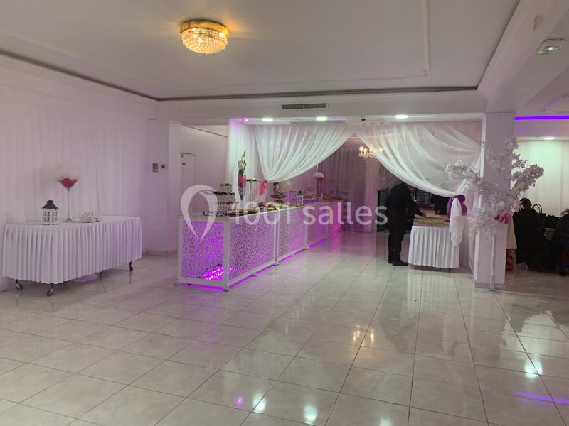 Salle de réception lumineuse avec tables décorées, buffet éclairé en rose et rideaux blancs suspendus.