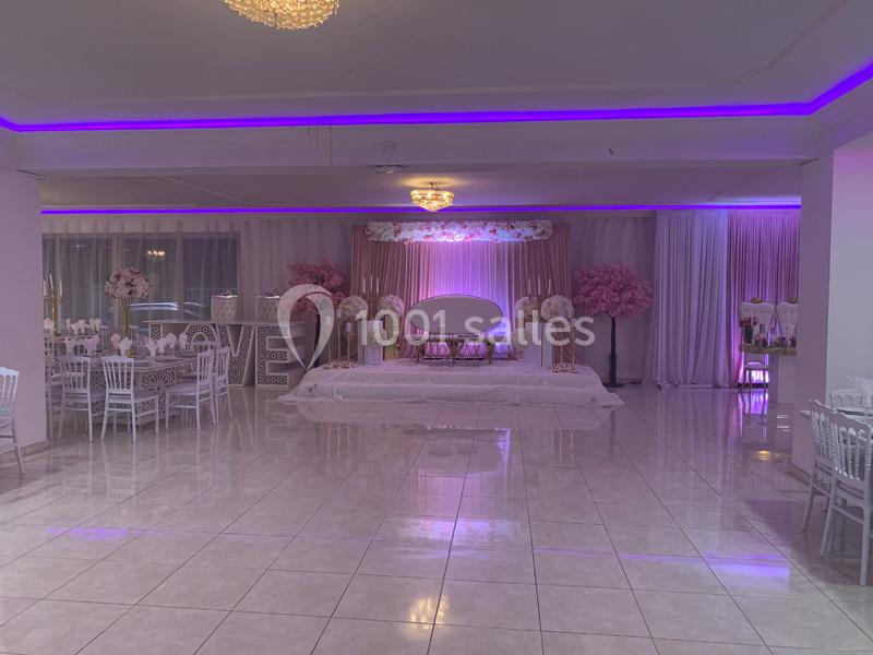 Salle de réception décorée avec des tons roses et violets, comprenant une scène centrale et des tables élégamment dressées.