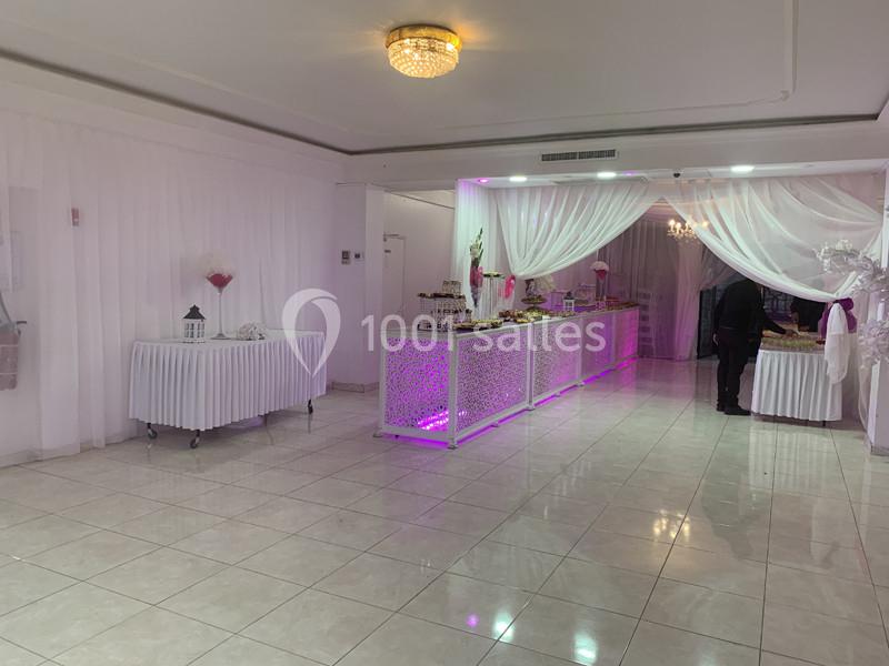 Salle de réception lumineuse avec tables décorées, nappes blanches et éclairage rose près d'un buffet.