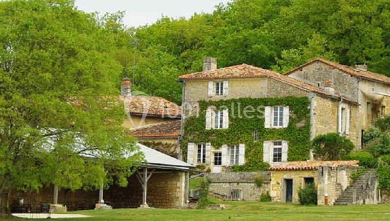 Maison en pierre recouverte de lierre, entourée d'arbres et d'une annexe avec toit en tuiles, dans un cadre rural.