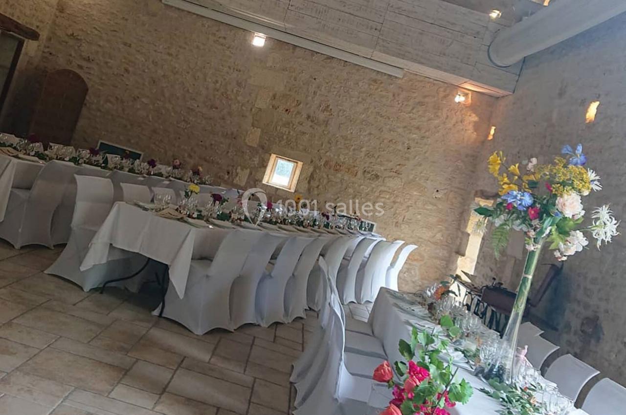 Salle en pierre aménagée pour un événement avec de longues tables décorées de nappes blanches et de fleurs colorées.