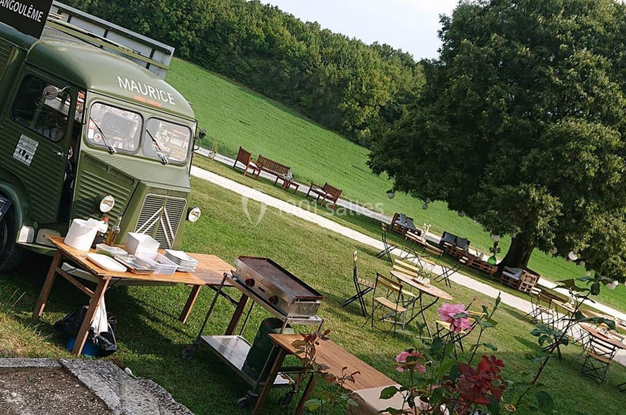 Camion-restaurant vert stationné dans un parc avec tables en bois, chaises et végétation environnante.