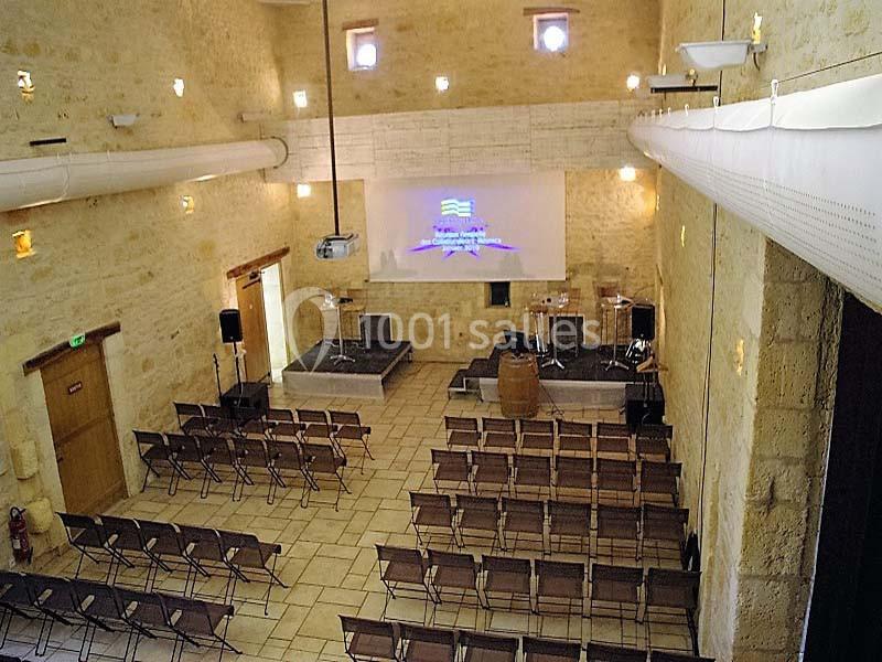 Salle en pierre avec des rangées de chaises, une scène équipée de micros et un écran de projection allumé.