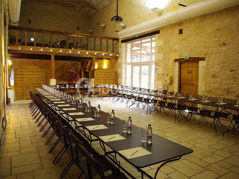 Salle en pierre avec de longues tables alignées, préparées avec des bouteilles d'eau, des verres et des dossiers.