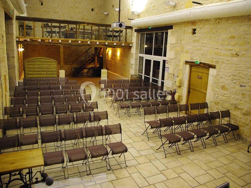 Salle en pierre avec des rangées de chaises alignées, un projecteur au plafond et une mezzanine en bois.