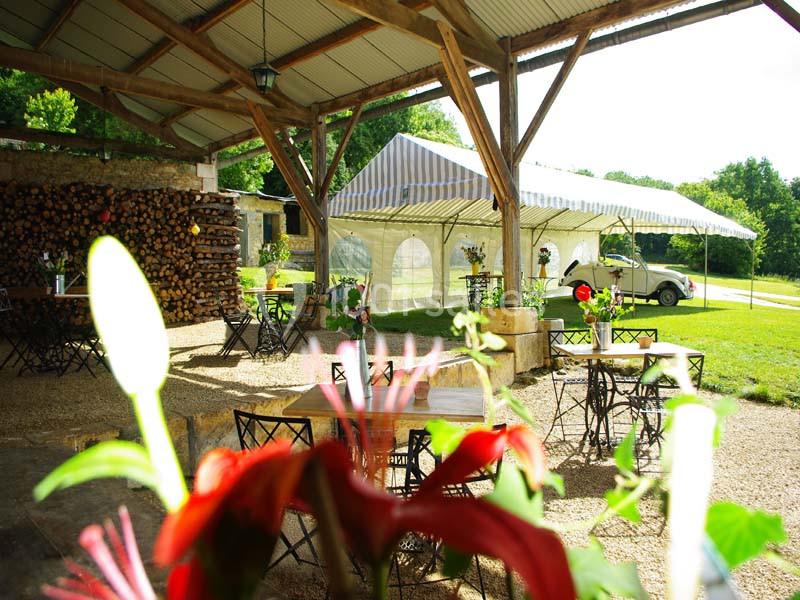 Espace extérieur avec tables et chaises sous une structure en bois, tente blanche en arrière-plan et verdure environnante.