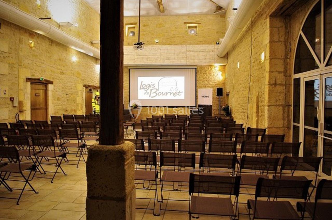 Salle de conférence en pierre avec des rangées de chaises pliantes et un écran de projection allumé au fond.