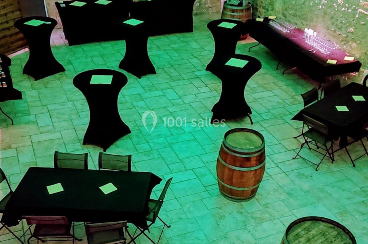 Salle aménagée pour un événement, avec tables hautes et basses noires, tonneaux en bois et éclairage vert.