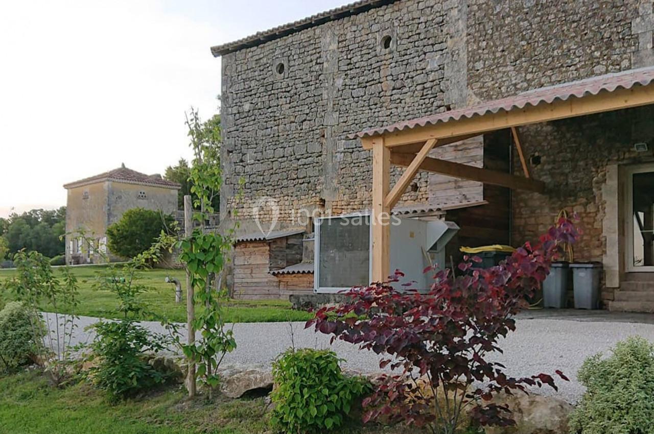 Bâtiments en pierre dans un cadre rural avec un espace vert, une pergola en bois et des arbustes au premier plan.
