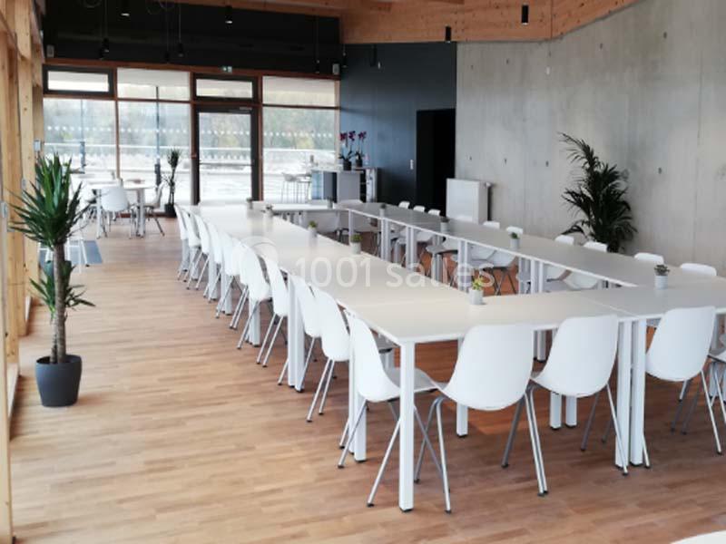 Salle lumineuse avec de grandes baies vitrées, tables blanches alignées en U et chaises modernes sur un sol en bois.