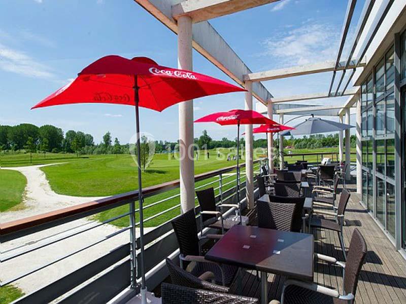 Terrasse avec tables, chaises en osier et parasols rouges, surplombant un terrain de golf par temps ensoleillé.