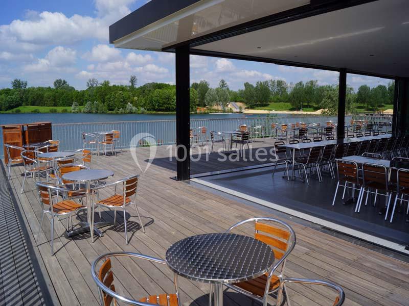 Terrasse en bois avec tables et chaises en métal, surplombant un lac entouré de verdure par temps ensoleillé.