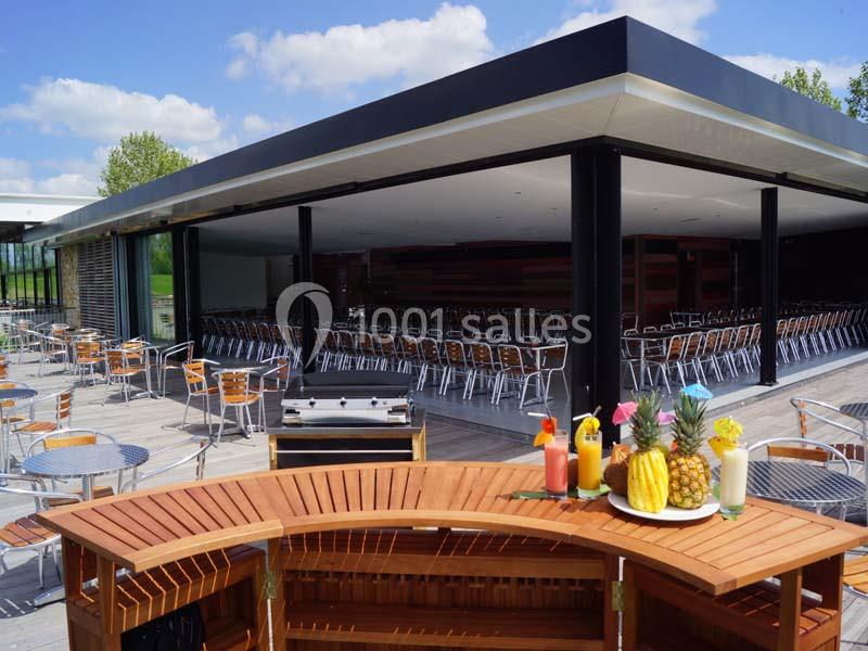 Terrasse avec tables et chaises en métal, bar en bois au premier plan et salle intérieure avec sièges alignés.