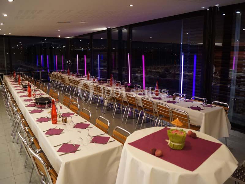 Salle de réception avec tables alignées, nappes blanches, serviettes violettes et éclairage coloré en arrière-plan.