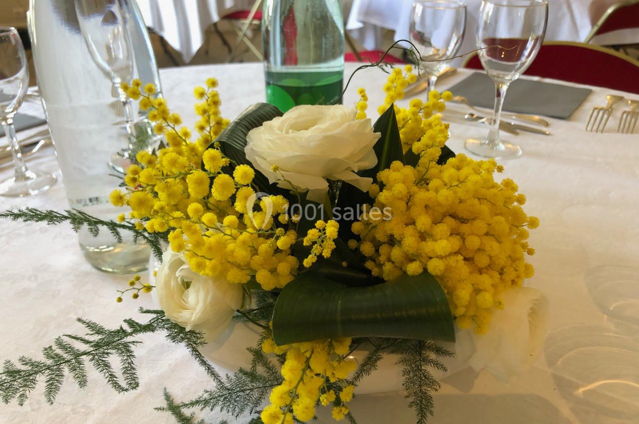 Centre de table avec des fleurs jaunes de mimosa, des roses blanches et des feuilles vertes sur une nappe blanche.