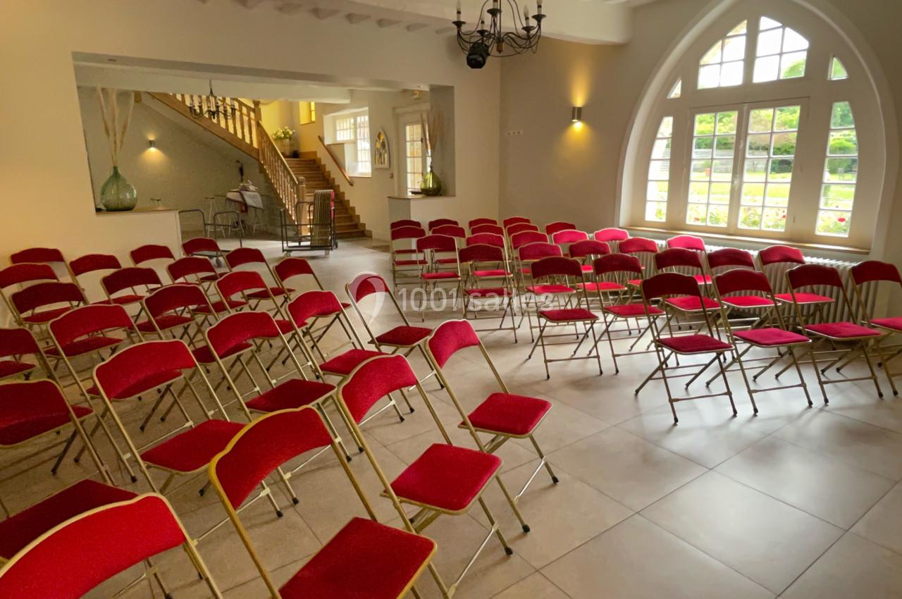 Salle lumineuse avec des chaises rouges alignées, un escalier en bois et de grandes fenêtres donnant sur un jardin.