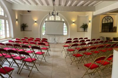 Salle lumineuse avec une grande table rectangulaire couverte d'une nappe bleue, entourée de chaises rouges, et une fenêtre…