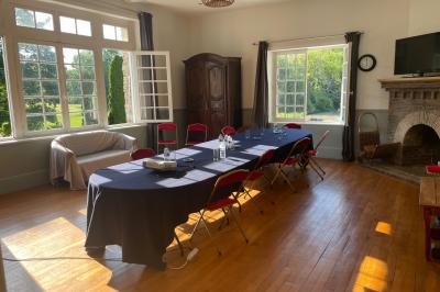 Salle lumineuse avec une grande table rectangulaire couverte d'une nappe bleue, entourée de chaises rouges, et une fenêtre…