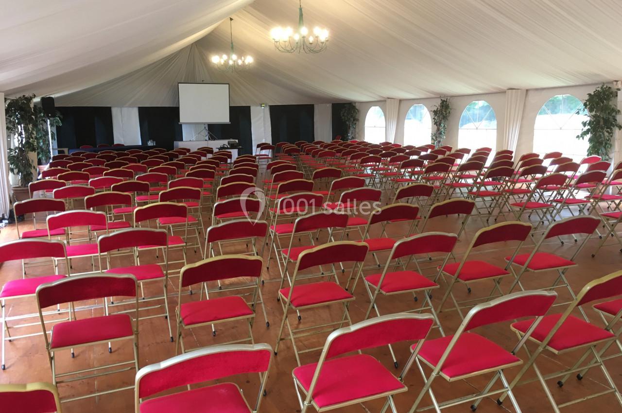 Salle de conférence sous une tente avec rangées de chaises rouges alignées face à un écran de projection.