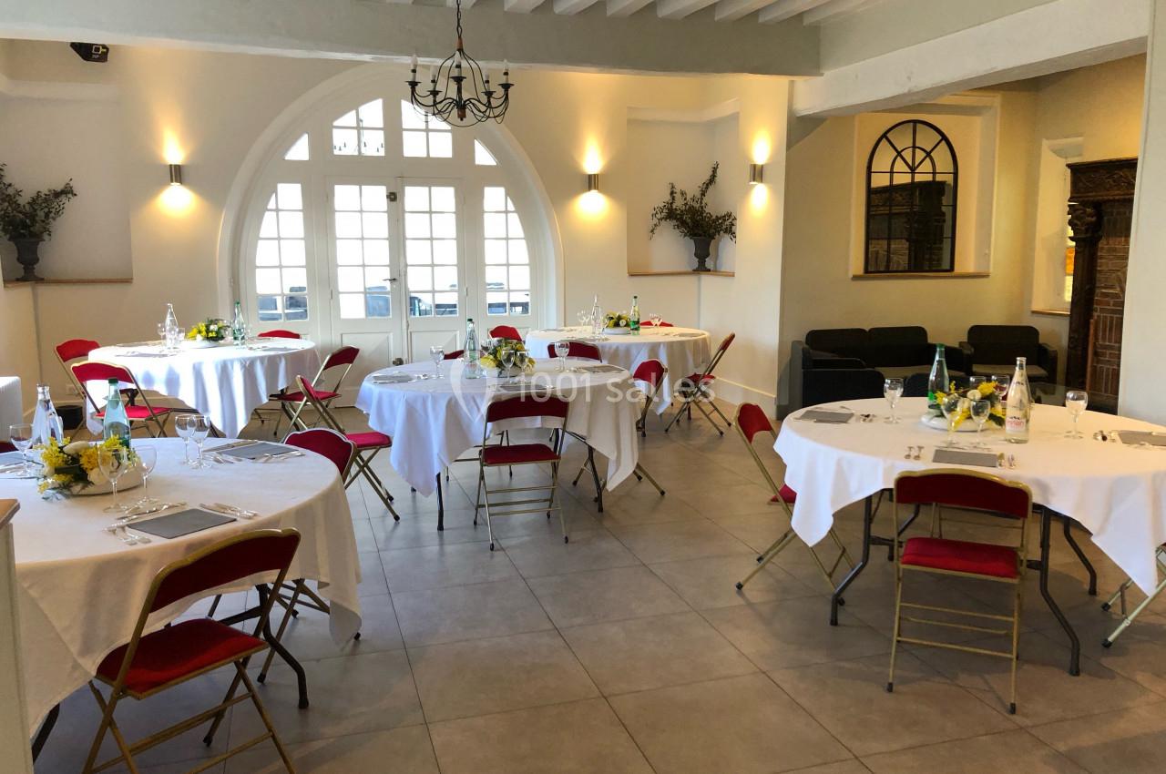 Salle lumineuse avec tables rondes dressées, chaises rouges, grandes fenêtres et décoration sobre.