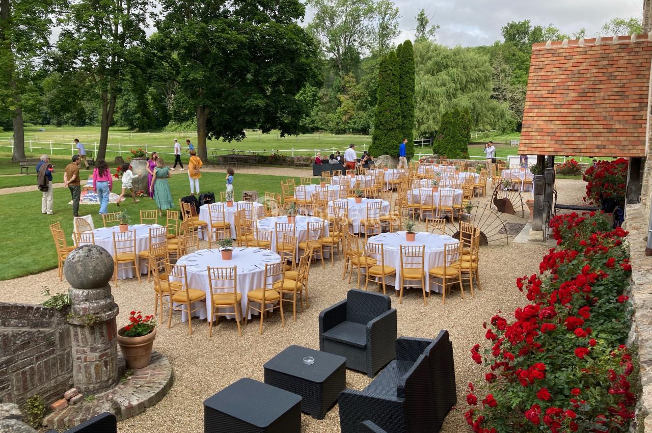 Tables rondes dressées avec nappes blanches et chaises en bois disposées en extérieur dans un jardin fleuri.