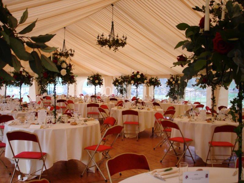 Salle de réception sous une tente avec tables rondes dressées, chaises rouges et décorations florales suspendues.