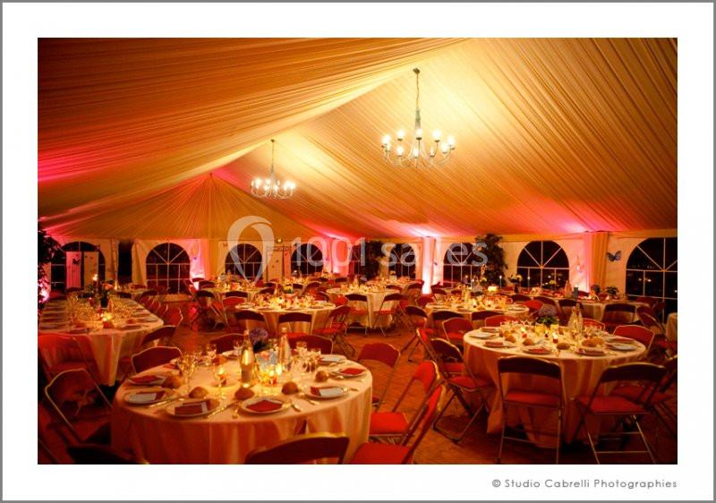 Salle de réception sous une tente avec tables rondes dressées, éclairage chaleureux et décorations élégantes.