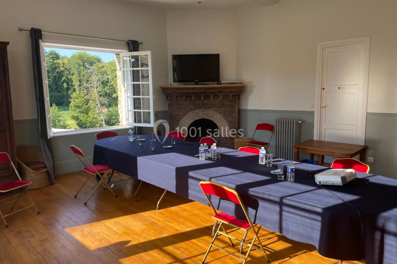 Salle lumineuse avec une grande table rectangulaire couverte d'une nappe bleue, entourée de chaises rouges, et une fenêtre…
