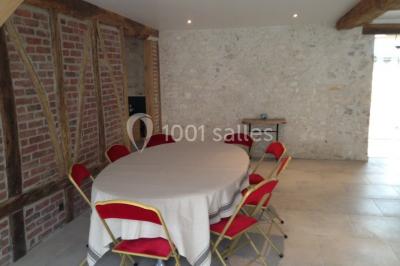 Salle lumineuse avec une grande table rectangulaire couverte d'une nappe bleue, entourée de chaises rouges, et une fenêtre…