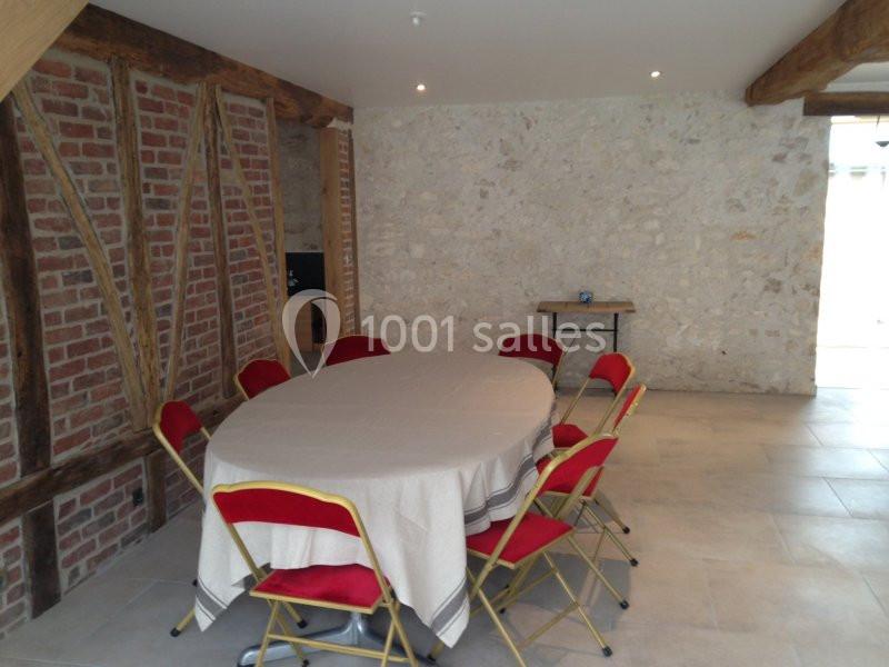 Salle avec murs en pierre et briques apparentes, table ronde couverte d'une nappe blanche entourée de chaises rouges.