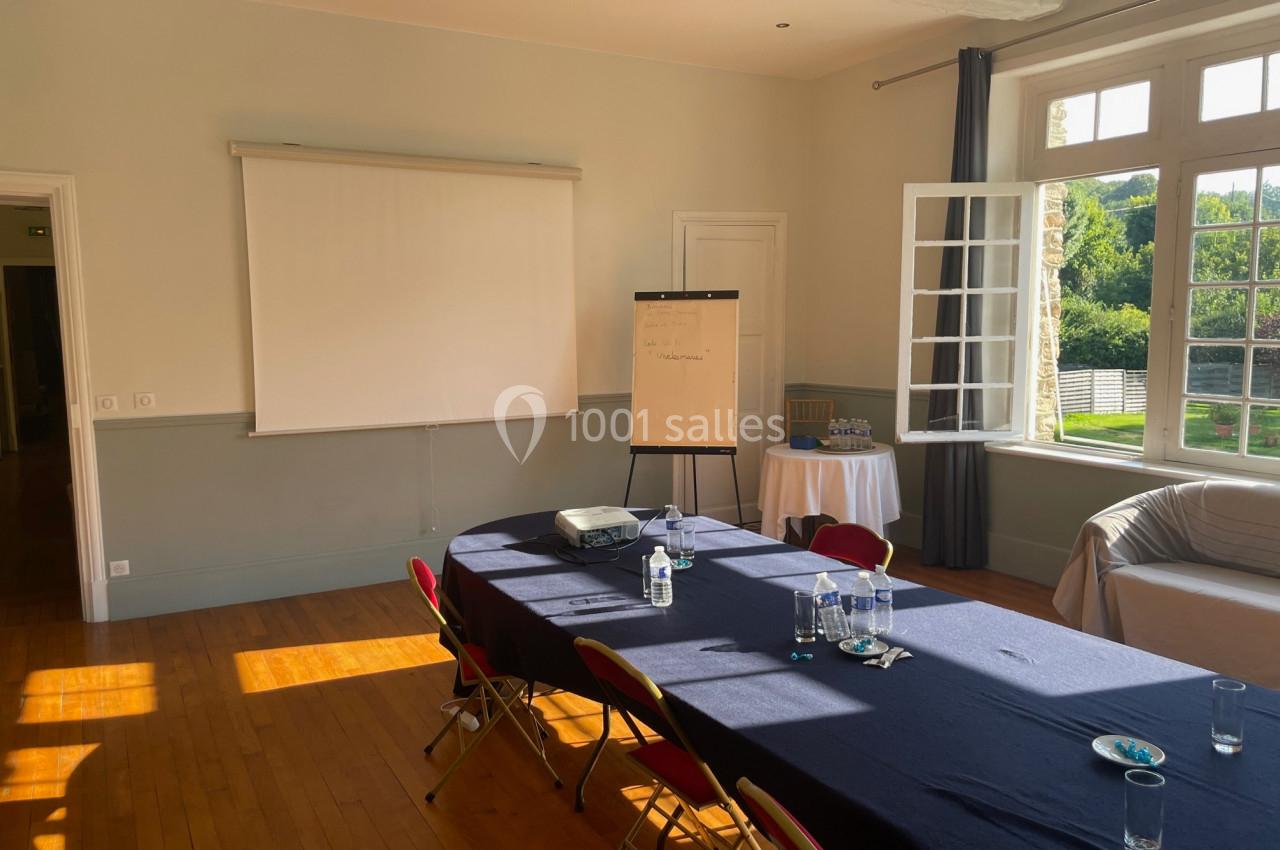 Salle de réunion lumineuse avec table, chaises, projecteur, écran, paperboard et vue sur un jardin par une fenêtre ouverte.