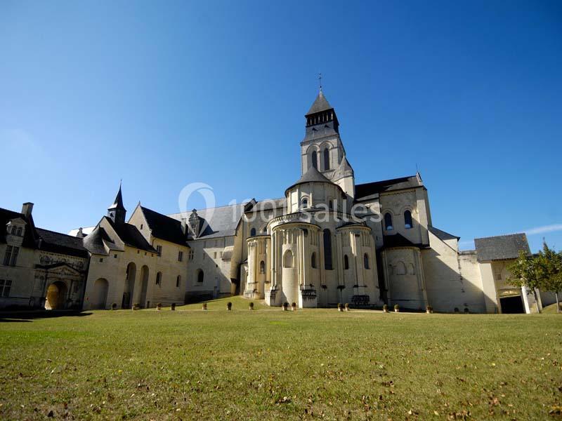 Abbaye médiévale avec clocher et bâtiments en pierre, entourée d'une pelouse sous un ciel bleu clair.