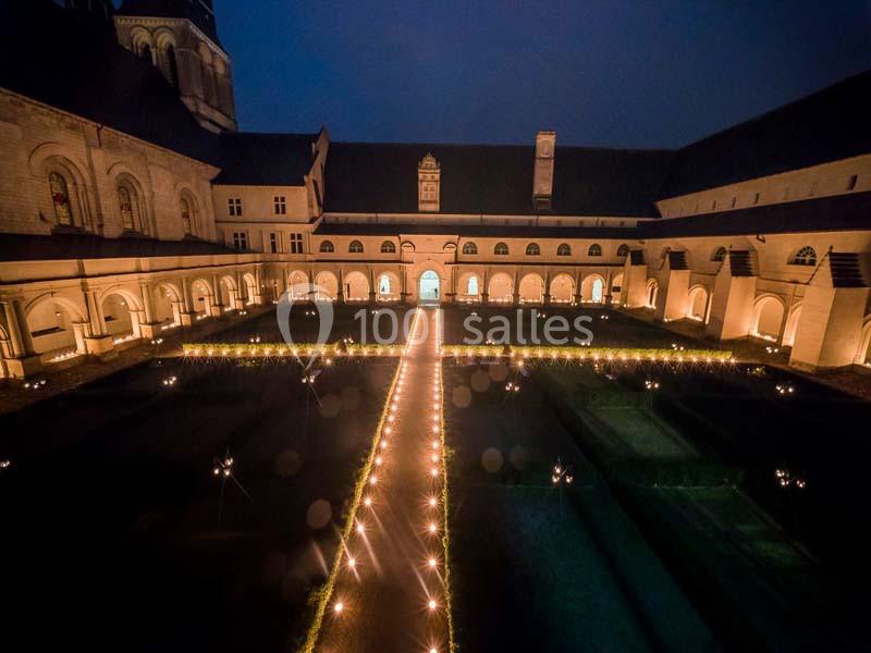 Cloître illuminé de nuit avec des allées éclairées formant une croix au centre, entouré d'arcades.
